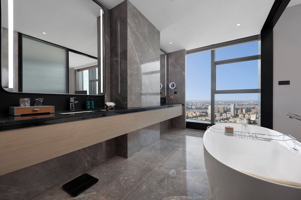 A modern bathroom with a large bathtub, a vanity with a mirror, and a view of a cityscape through large windows.