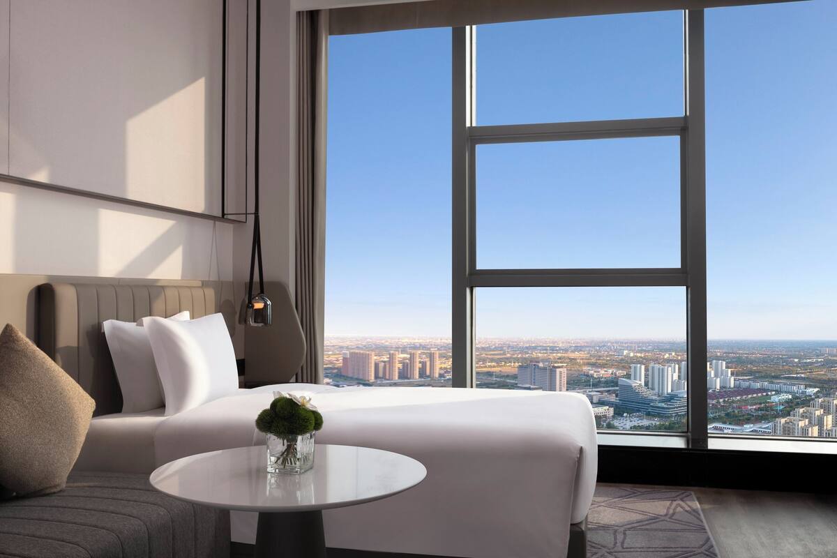 A modern hotel room with a large window offering a city view, a neatly made bed, a round table with a vase, and a sofa.