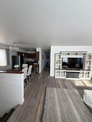 Een moderne woonkamer met een flatscreen televisie, een eethoek met een houten tafel, en een keuken op de achtergrond.