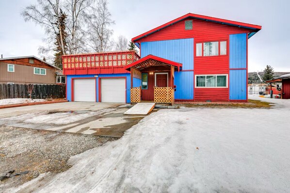 Uma casa de dois andares com fachada em vermelho e azul, varanda frontal e duas portas de garagem.