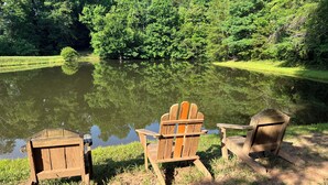A serene lakeside setting with three Adirondack chairs facing a calm pond surrounded by lush greenery. - The Cottage at Cardinal Hill (Dahlonega)