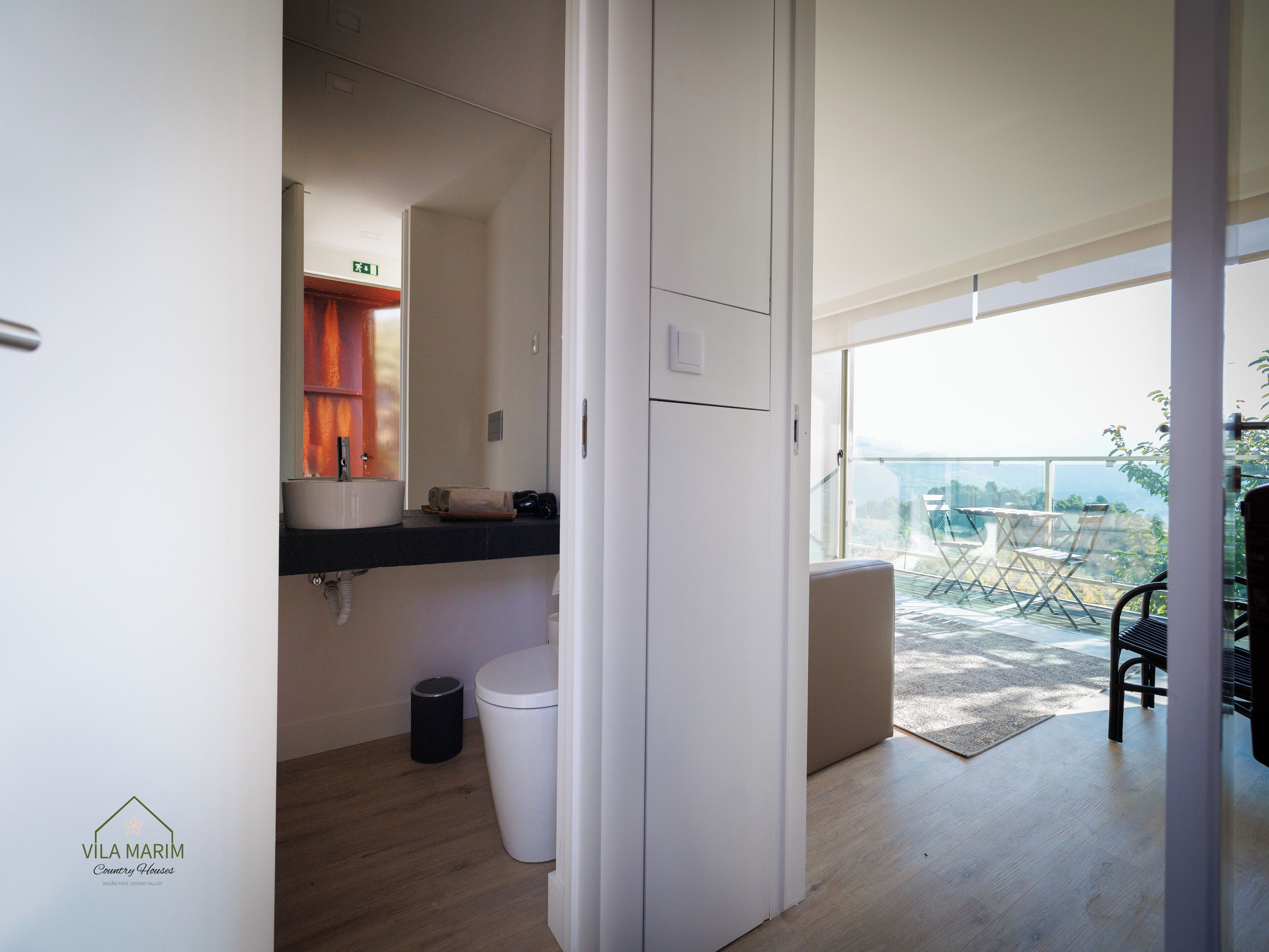 Ein modernes Badezimmer mit Waschbecken, Spiegel und Toilette. Ein kleiner Mülleimer und eine Tür zu einem Balkon mit Blick auf die Stadt.