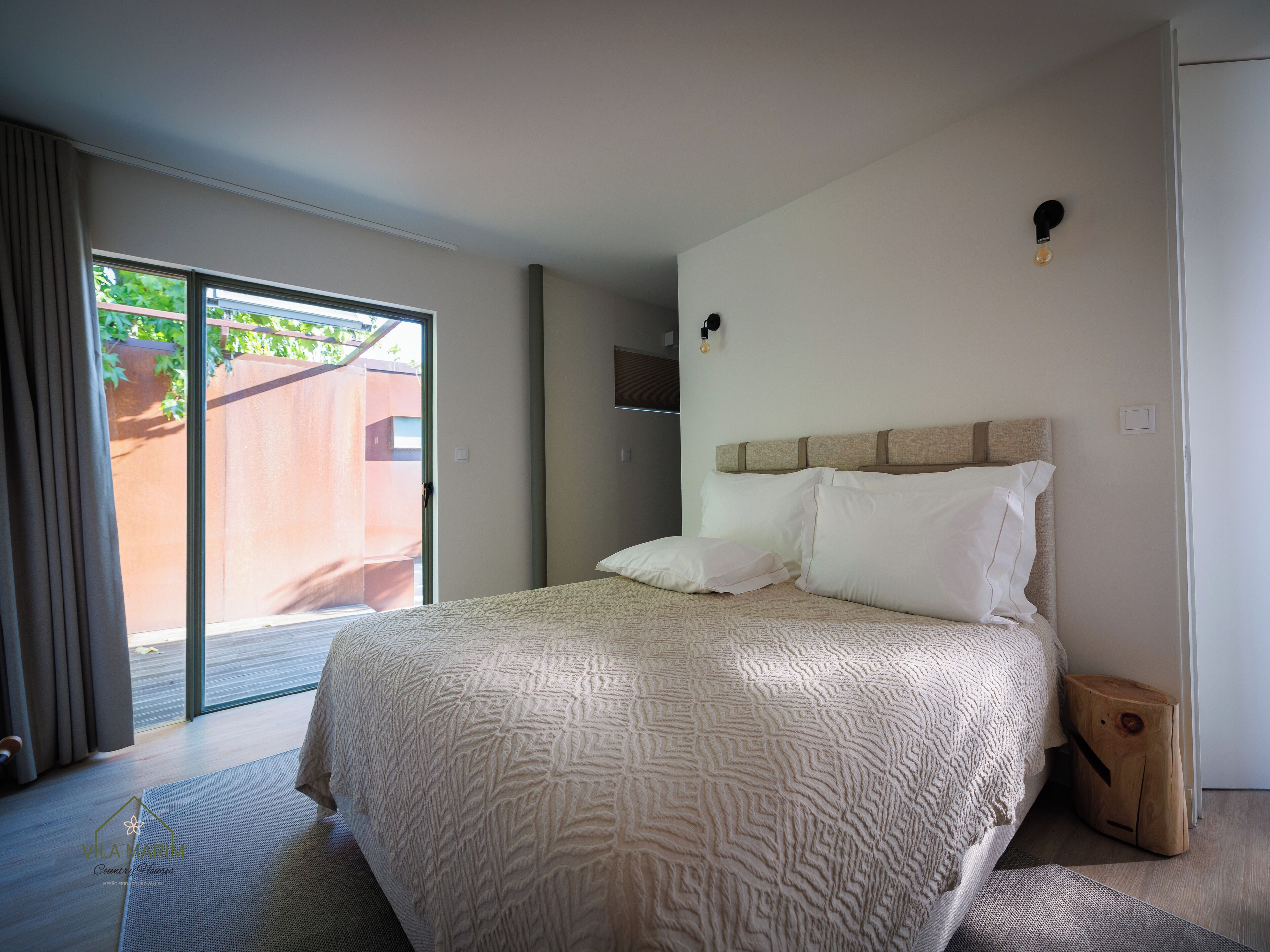 Quarto com uma cama grande, porta de correr de vidro para uma varanda e uma pequena mesa de cabeceira de madeira.