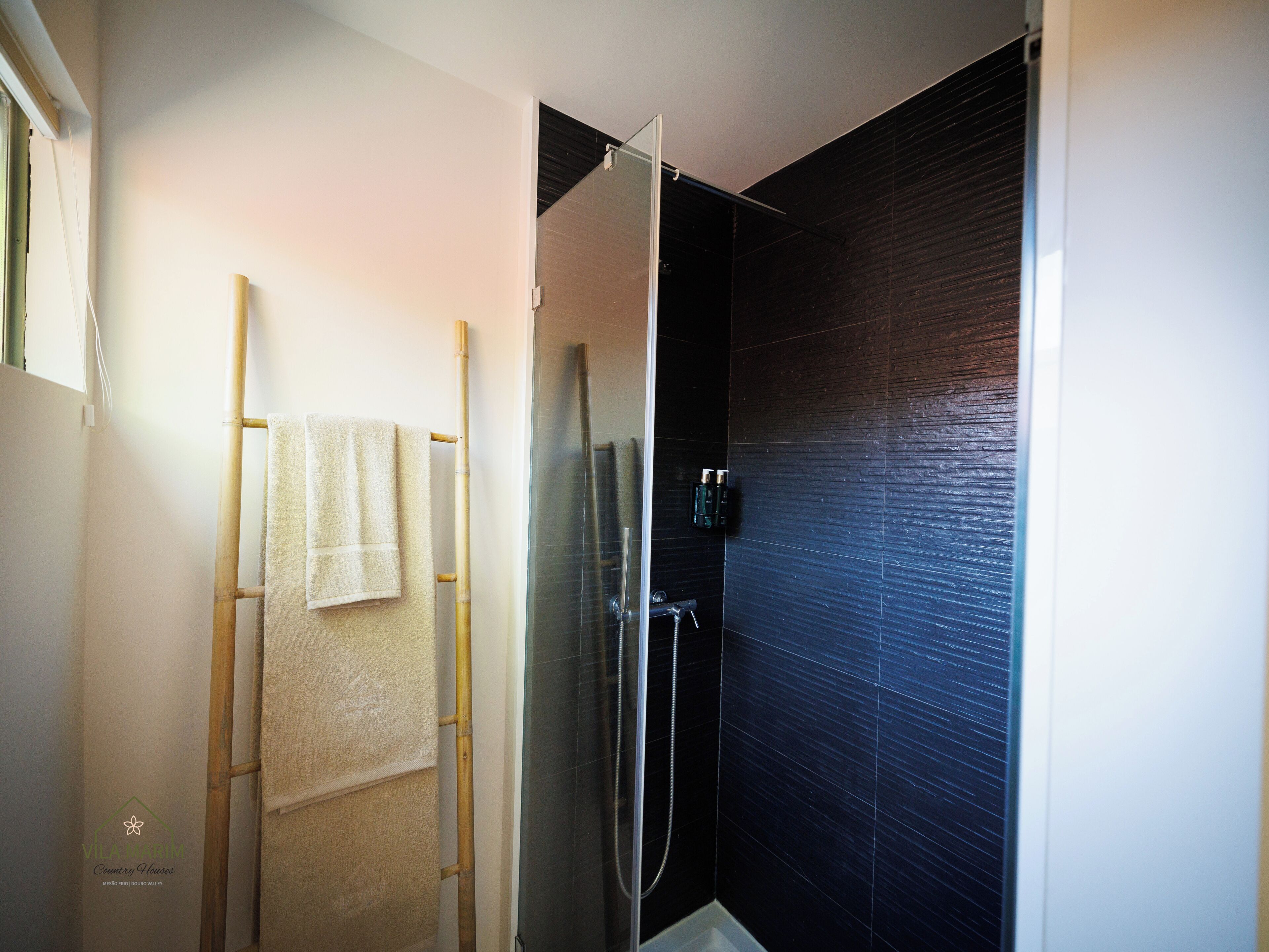 Une salle de bain dotée d’une douche à parois de verre, d’un porte-serviettes en bambou et d’un mur recouvert de carreaux foncés.