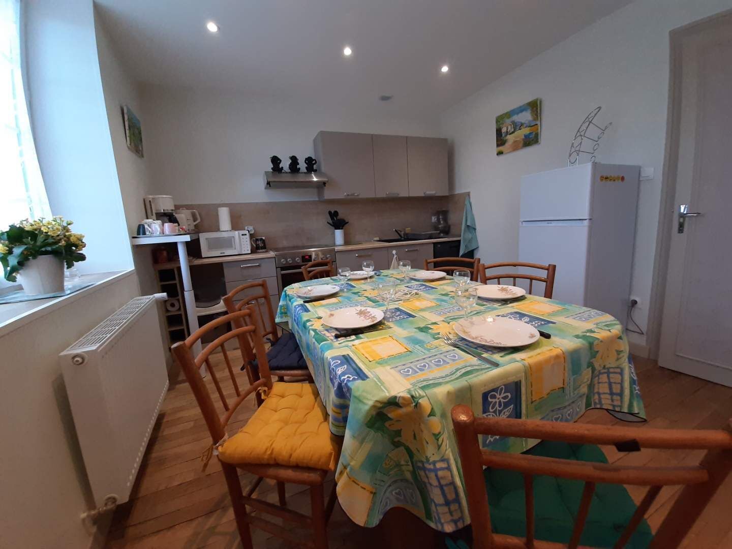 Un coin repas avec une table dressée pour quatre personnes, des chaises en bois, une cuisine en arrière-plan et un réfrigérateur sur la droite.