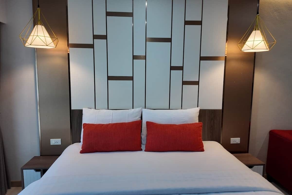 Habitación de hotel con cama, dos almohadas rojas, cabecera de madera y dos lámparas colgantes.