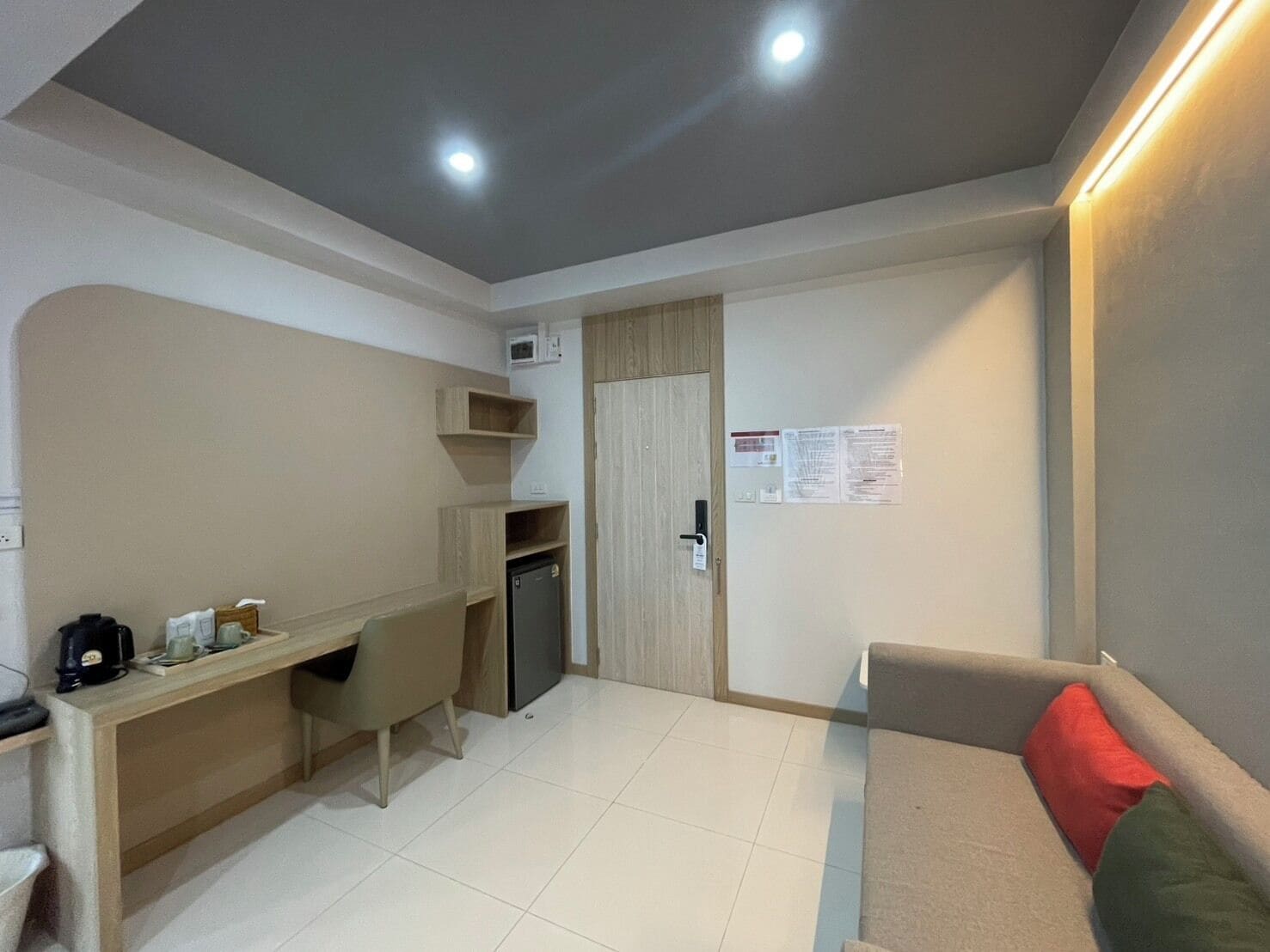 A compact room with a kitchenette, a desk, a chair, a small fridge, a sofa, and a door with a notice on the wall.