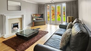 Una sala de estar moderna con chimenea, una mesa de centro de vidrio, un sofá y vistas a un jardín a través de amplios ventanales.