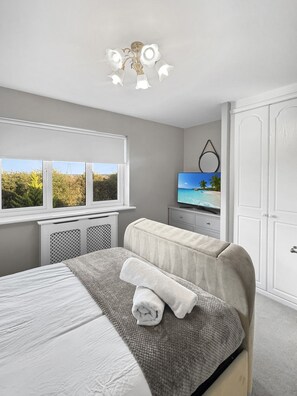 Un dormitorio con cama, televisor, ventana con vistas a zonas verdes y un armario blanco.