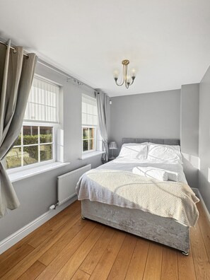 Ein Schlafzimmer mit einem Bett, zwei Fenstern mit Vorhängen, einem Heizkörper, einer Lampe und einer Chandelier.