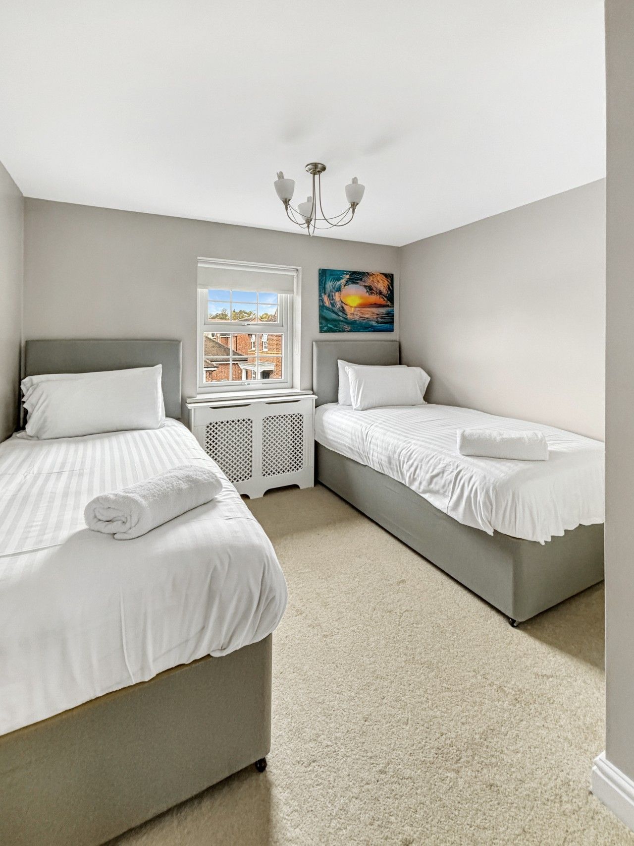 A bedroom with two beds, a window, a radiator, and a painting.