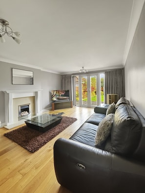 A modern living room with a fireplace, a glass coffee table, a leather sofa, and a view of a garden through large windows.