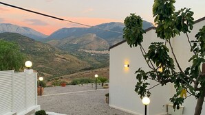 Un bâtiment blanc avec une allée de gravier et une vue sur les montagnes. On y trouve des plantes en pot et un arbre décoré de guirlandes lumineuses.