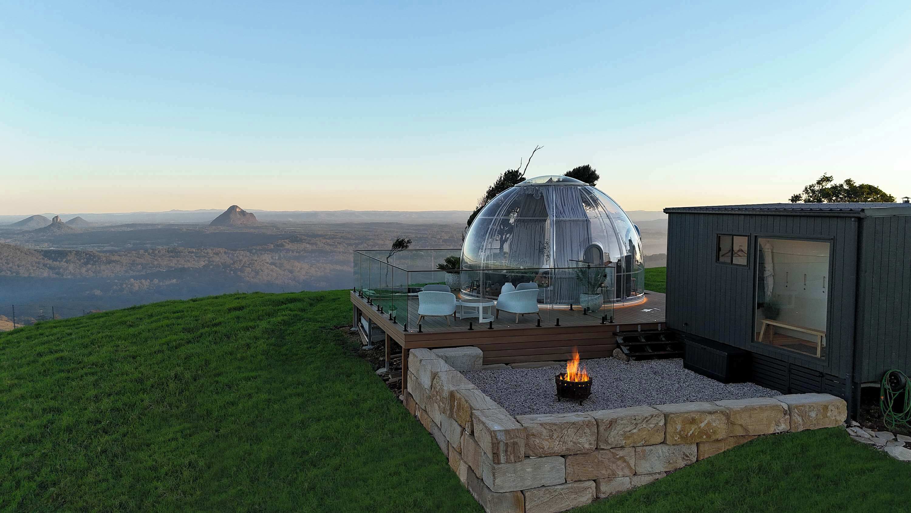 Eine gläserne Kuppelkonstruktion mit Feuerstelle, Sitzgelegenheiten im Freien und Blick auf ferne Hügel.