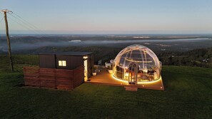Een koepelvormige glazen constructie met een houten terras en een klein aangrenzend gebouw.