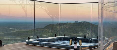 A hot tub with a glass enclosure, a bottle of wine, and a glass on a wooden deck with a scenic view.