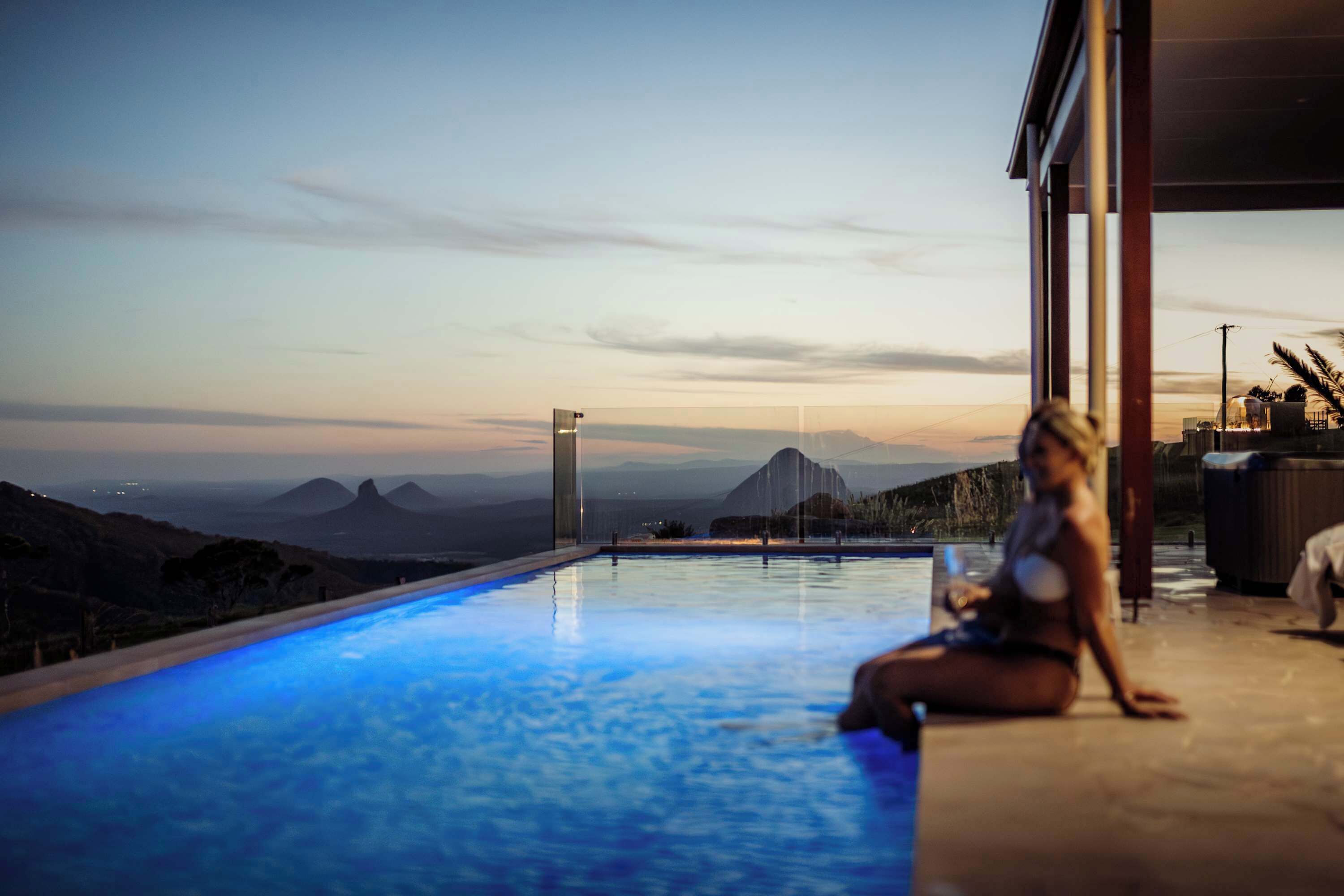 A person sitting by a pool with a view of mountains and a modern building in the background.