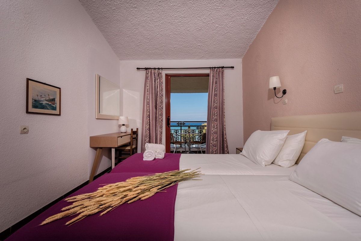 Ein Hotelzimmer mit Bett, Schreibtisch, Stuhl, einem Fenster mit Vorhängen, Meerblick und einem Gemälde an der Wand.