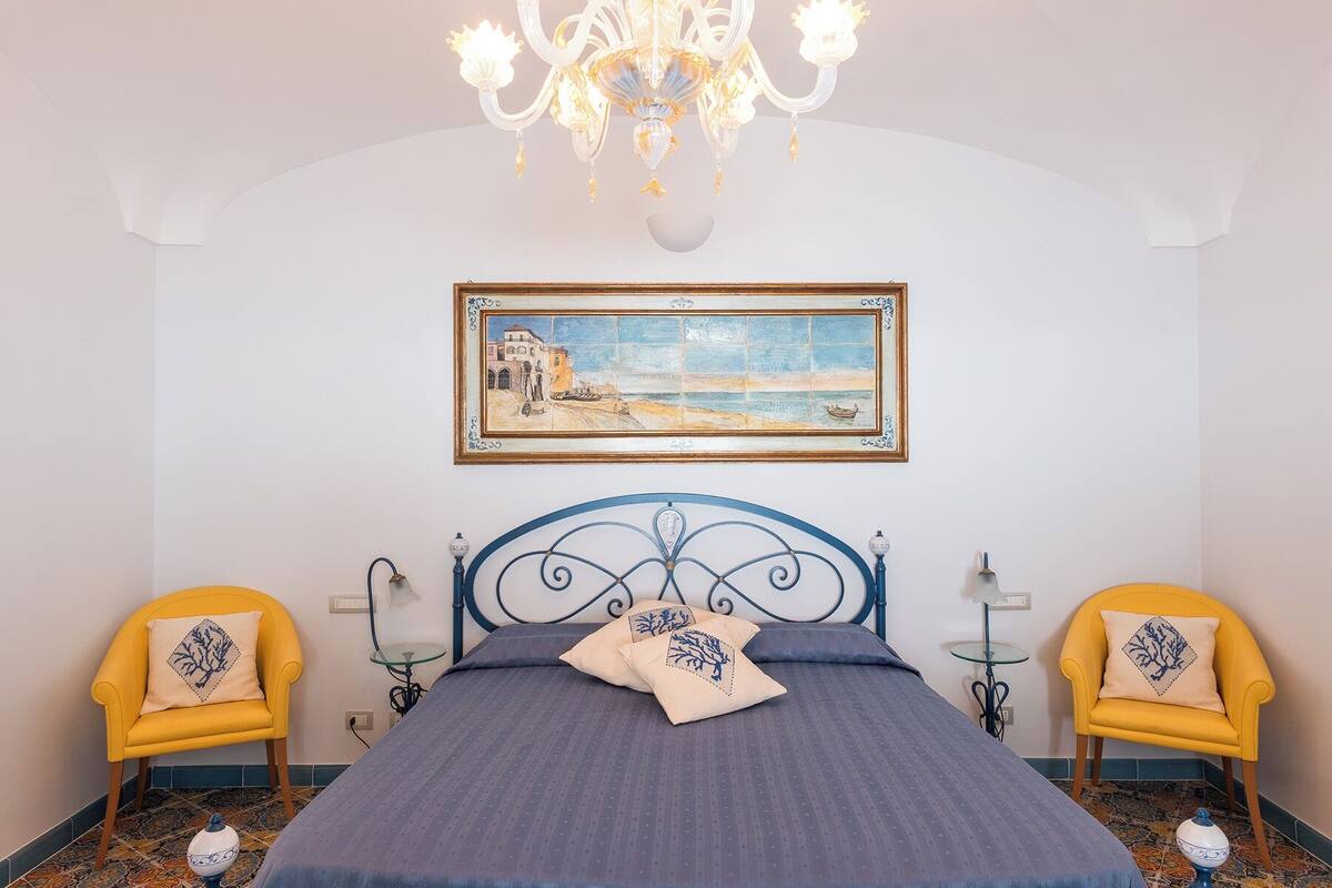A bedroom with a bed, two chairs, a chandelier, a painting, and a tiled floor.
