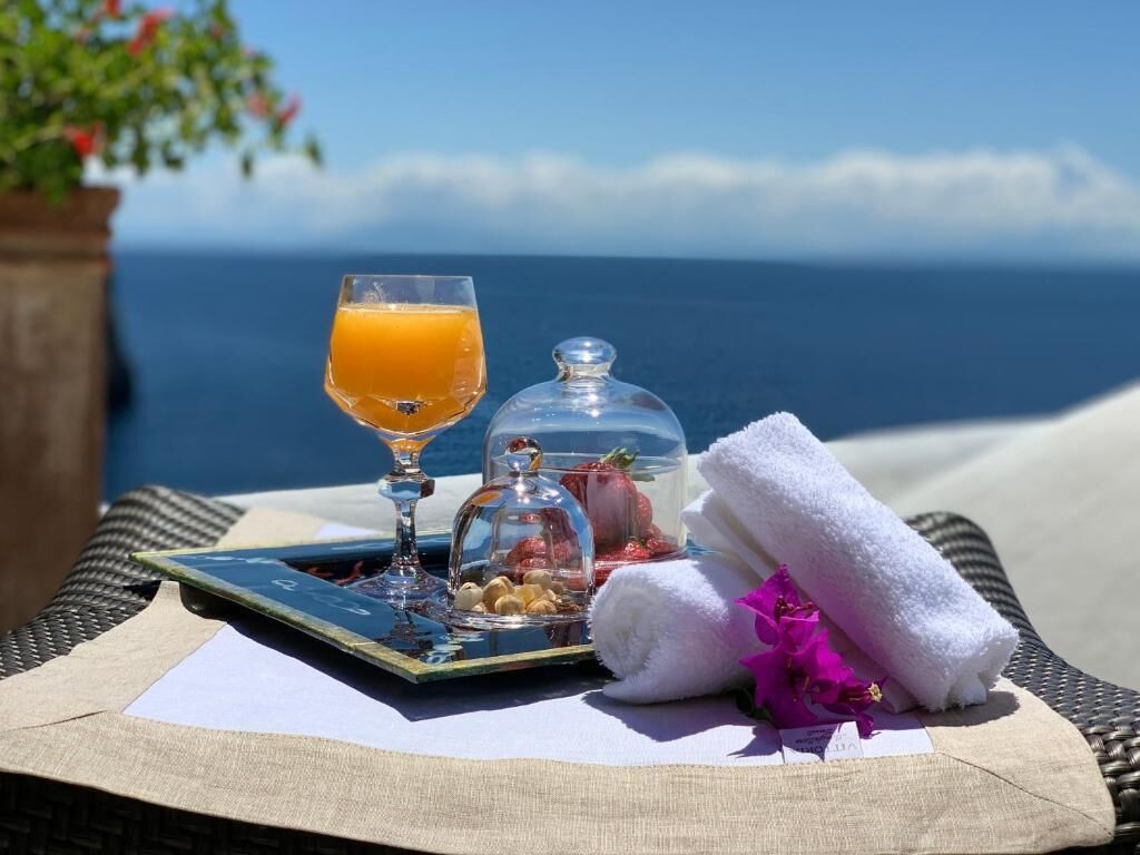 Ein Tablett mit einem Glas Orangensaft, einem Glas Wasser, einer Dose Nüsse, einer Dose Erdbeeren und einem gefalteten Handtuch auf einem Tisch mit Blick aufs Meer.