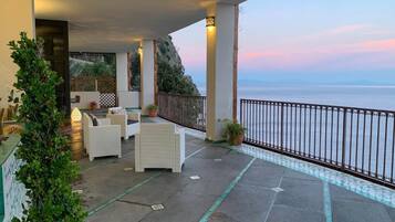 A covered outdoor seating area with a view of the sea, featuring a white wicker sofa and a small table.