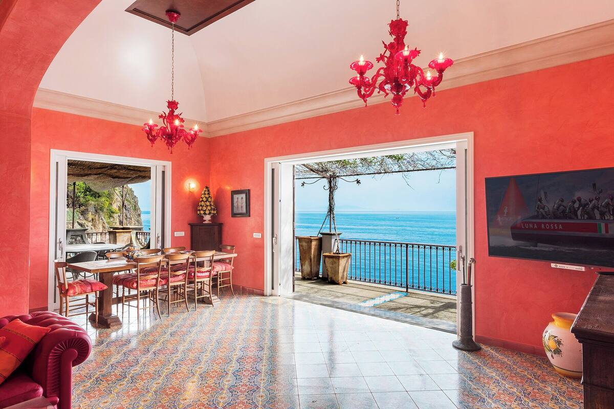 A room with a red wall, a dining area with chairs and a table, a chandelier, and a view of the sea through a balcony.