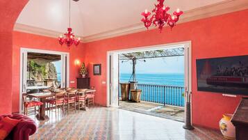 Ein Zimmer mit roter Wand, einem Essbereich mit Stühlen und Tisch, einer Kristallleuchtre und Meerblick vom Balkon.