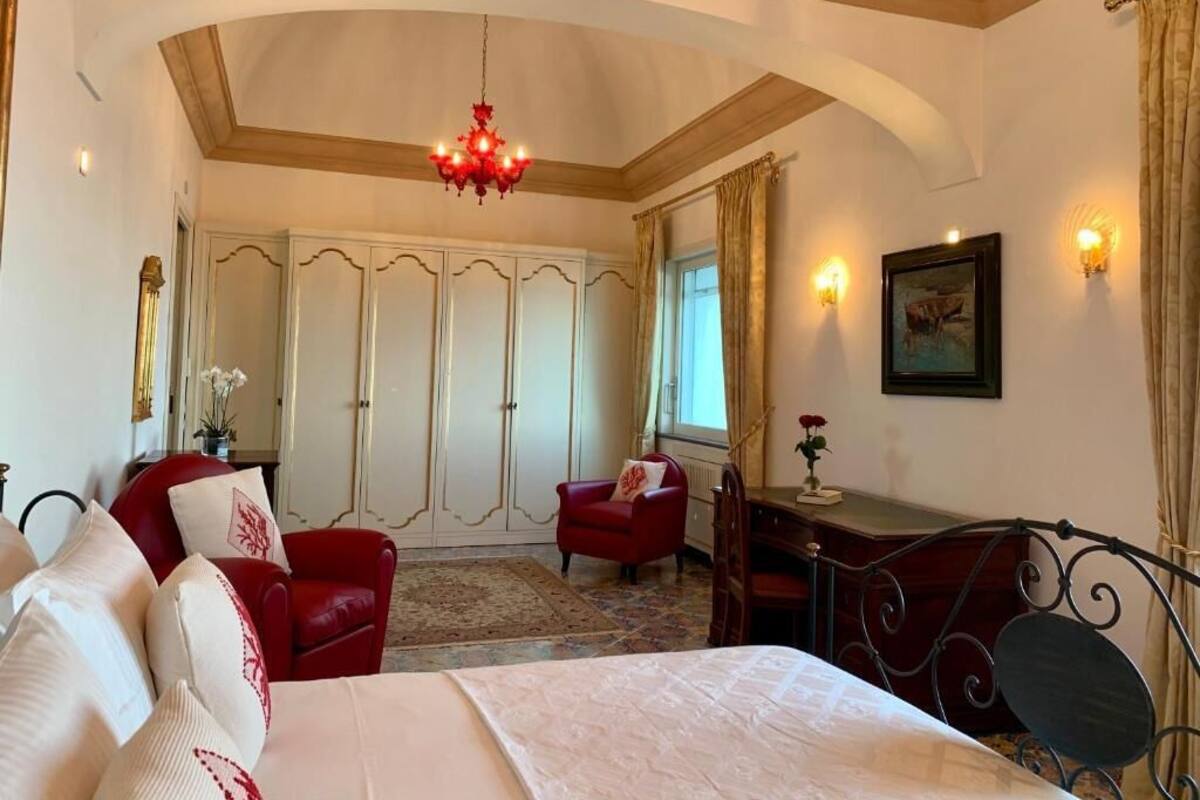 A bedroom with a bed, a red armchair, a desk, and a chandelier.