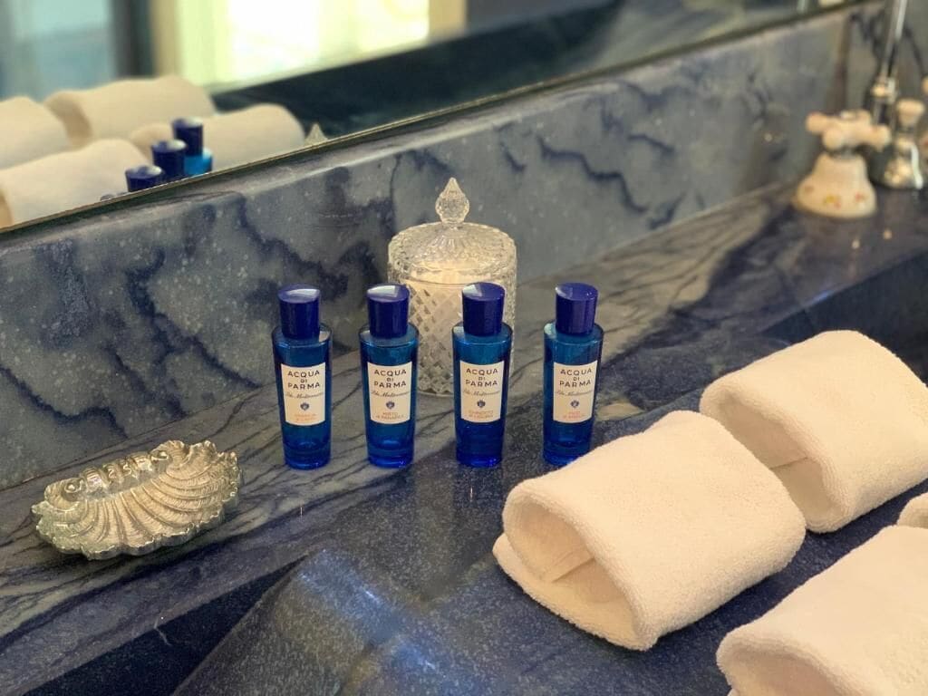 A bathroom vanity with a marble countertop, four blue bottles of Acqua Parma, a decorative shell, and two folded towels.