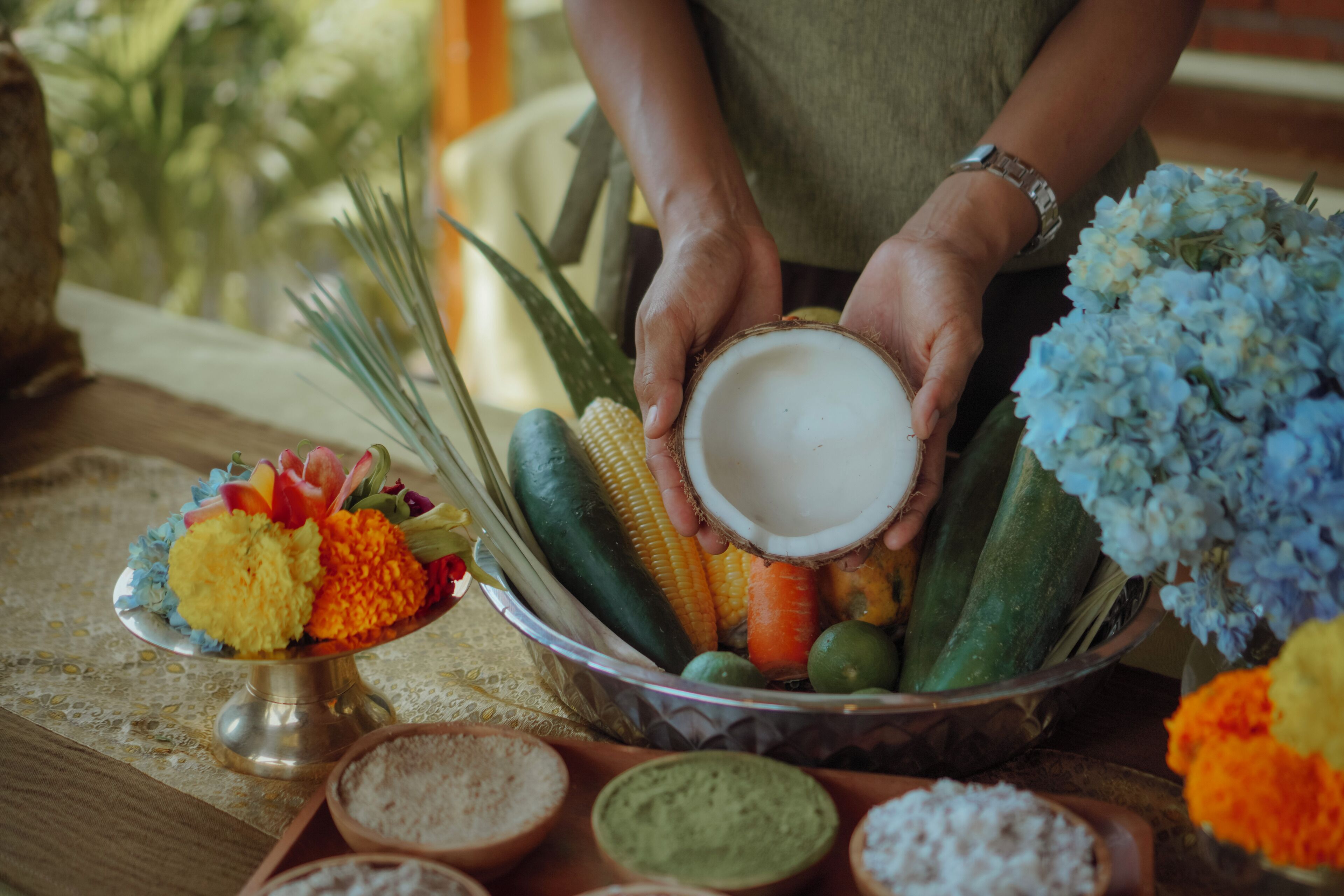 A person holding a coconut, surrounded by various vegetables and flowers on a table.