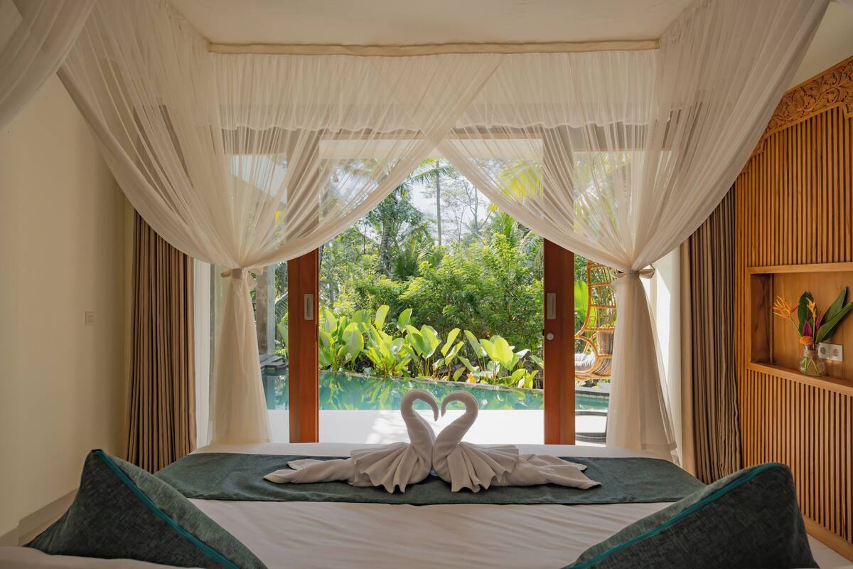 A bed with a canopy, a heart-shaped towel arrangement, and a view of a pool and greenery.