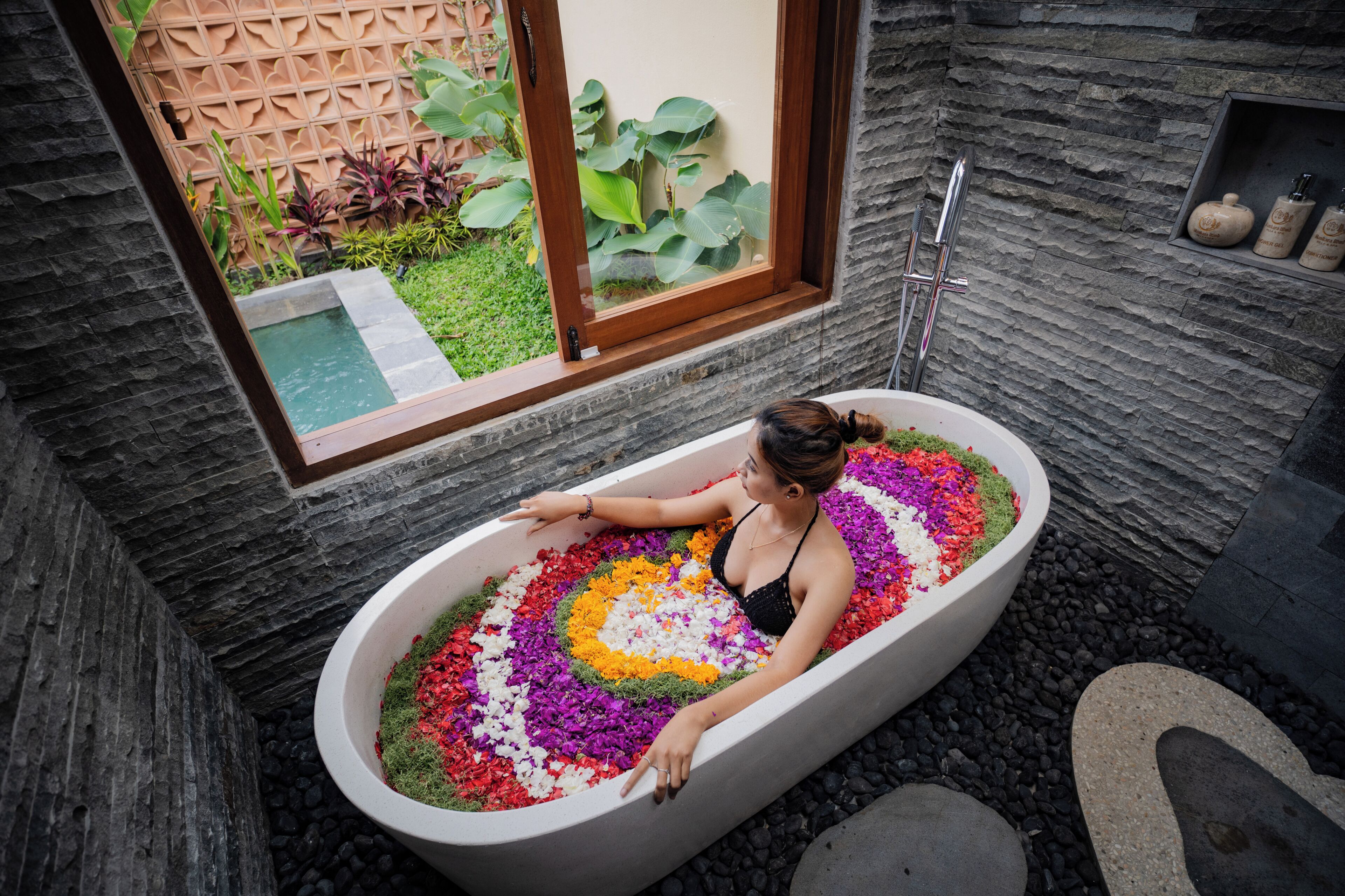 Una mujer en una bañera llena de pétalos de flores coloridos, con una ventana y plantas visibles al fondo.