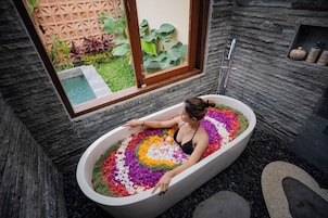 Una mujer en una bañera llena de pétalos de flores coloridos, con una ventana y plantas visibles al fondo.
