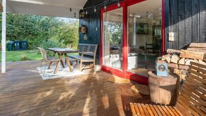 Una terraza de madera al aire libre con una mesa y sillas, una puerta de vidrio con marco rojo que da a una habitación, y una pila de leña.