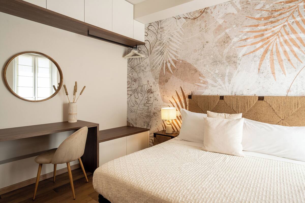 Een slaapkamer met een bed, een bureau, een stoel, een nachtkastje met een lamp en een muur met een bladmotief.