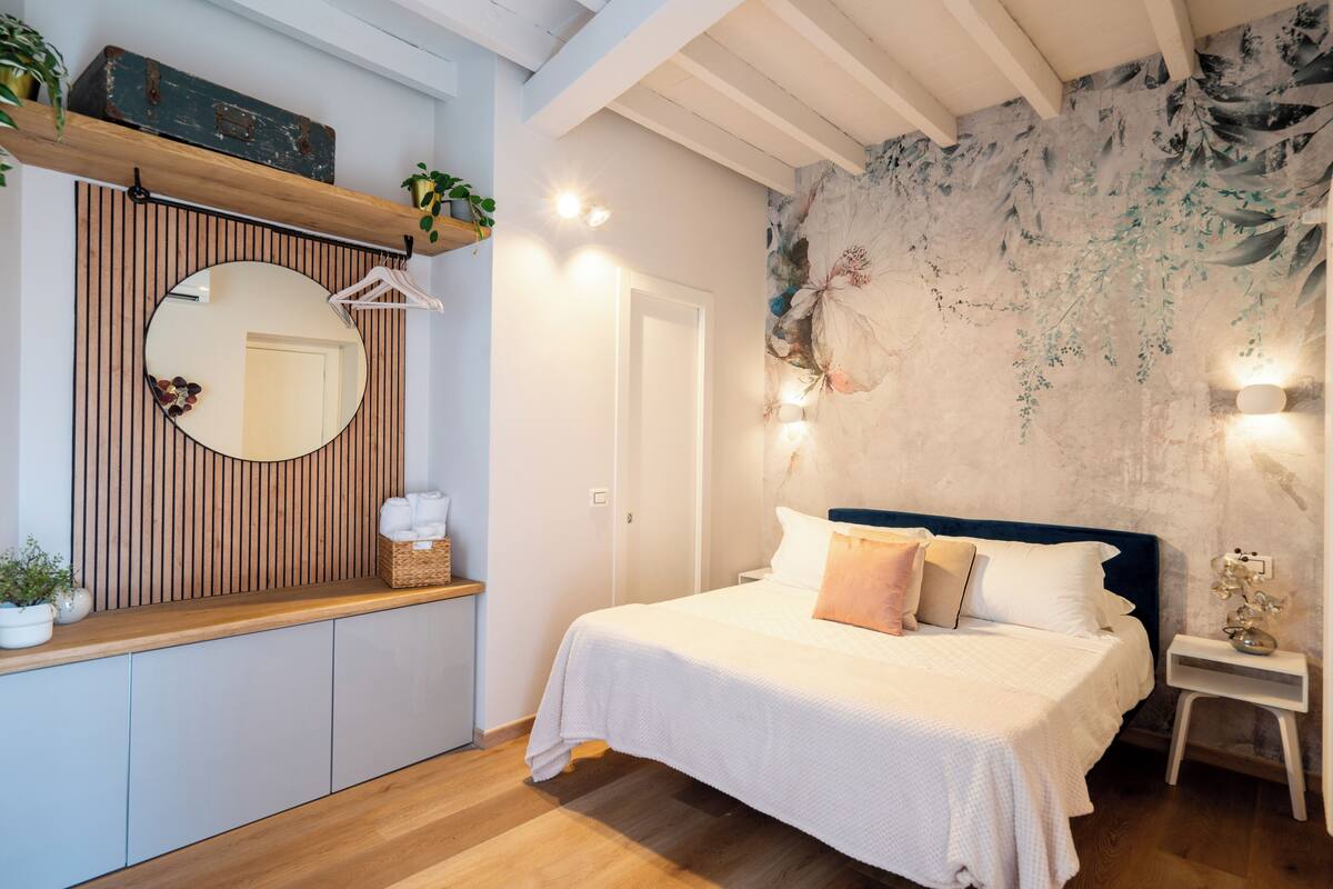 Een slaapkamer met een bed, een spiegel, een houten plank met planten en een muur met een bloemenmuur.