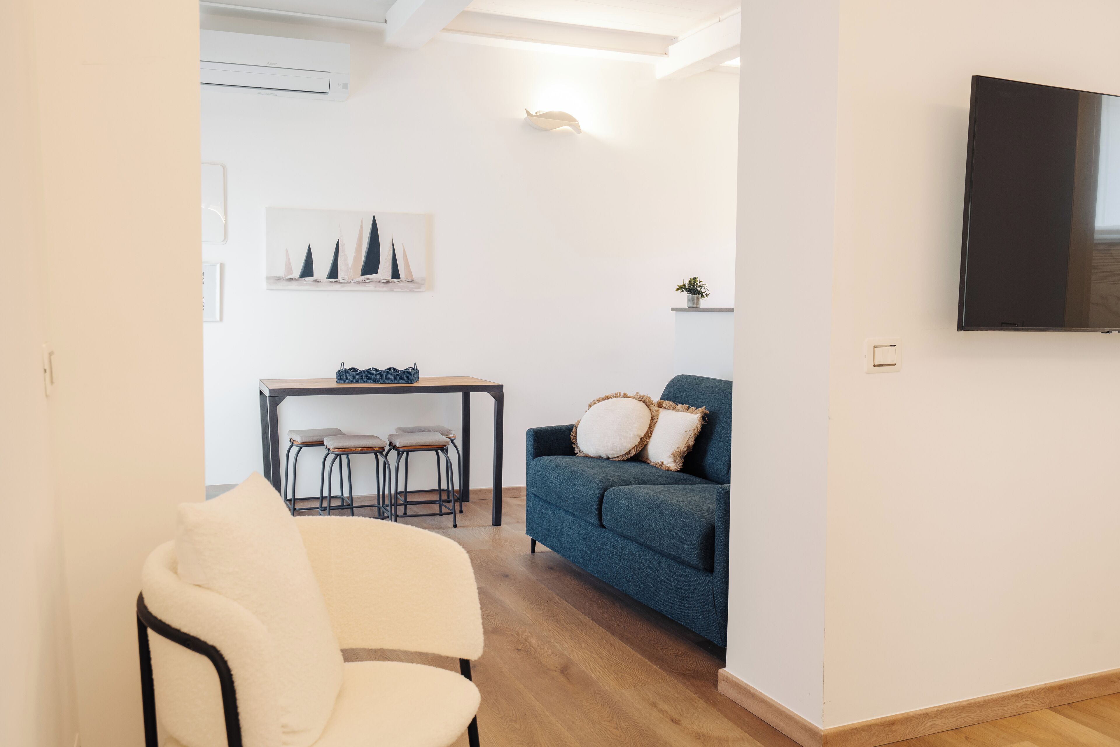 A modern living room with a sofa, a dining table with chairs, a TV mounted on the wall, and a small plant on a shelf.