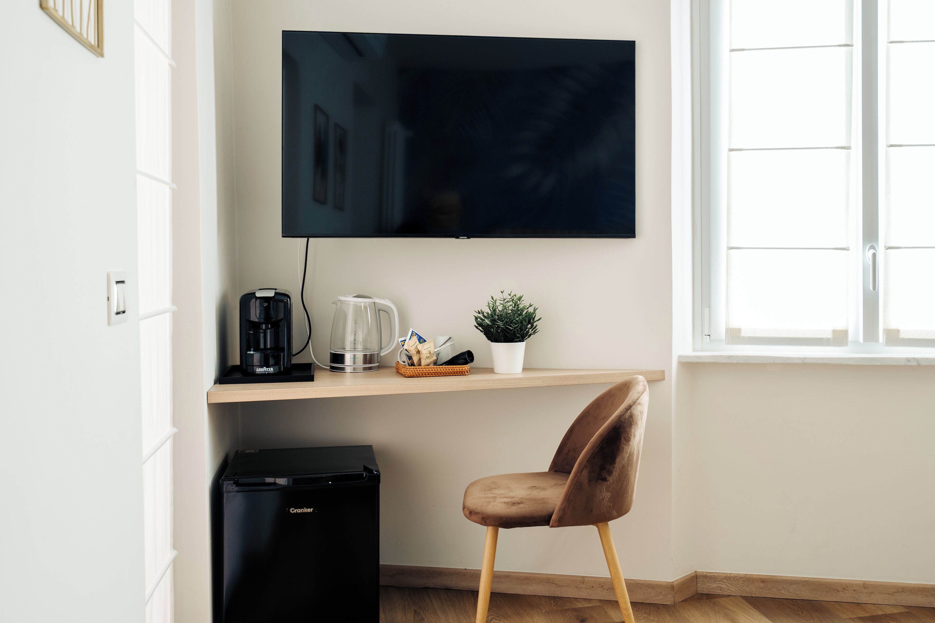 Een moderne kamer met een flatscreen televisie aan de muur, een kleine koelkast, een houten plankje met een koffiezetapparaat, een potplant en een bruine stoel.