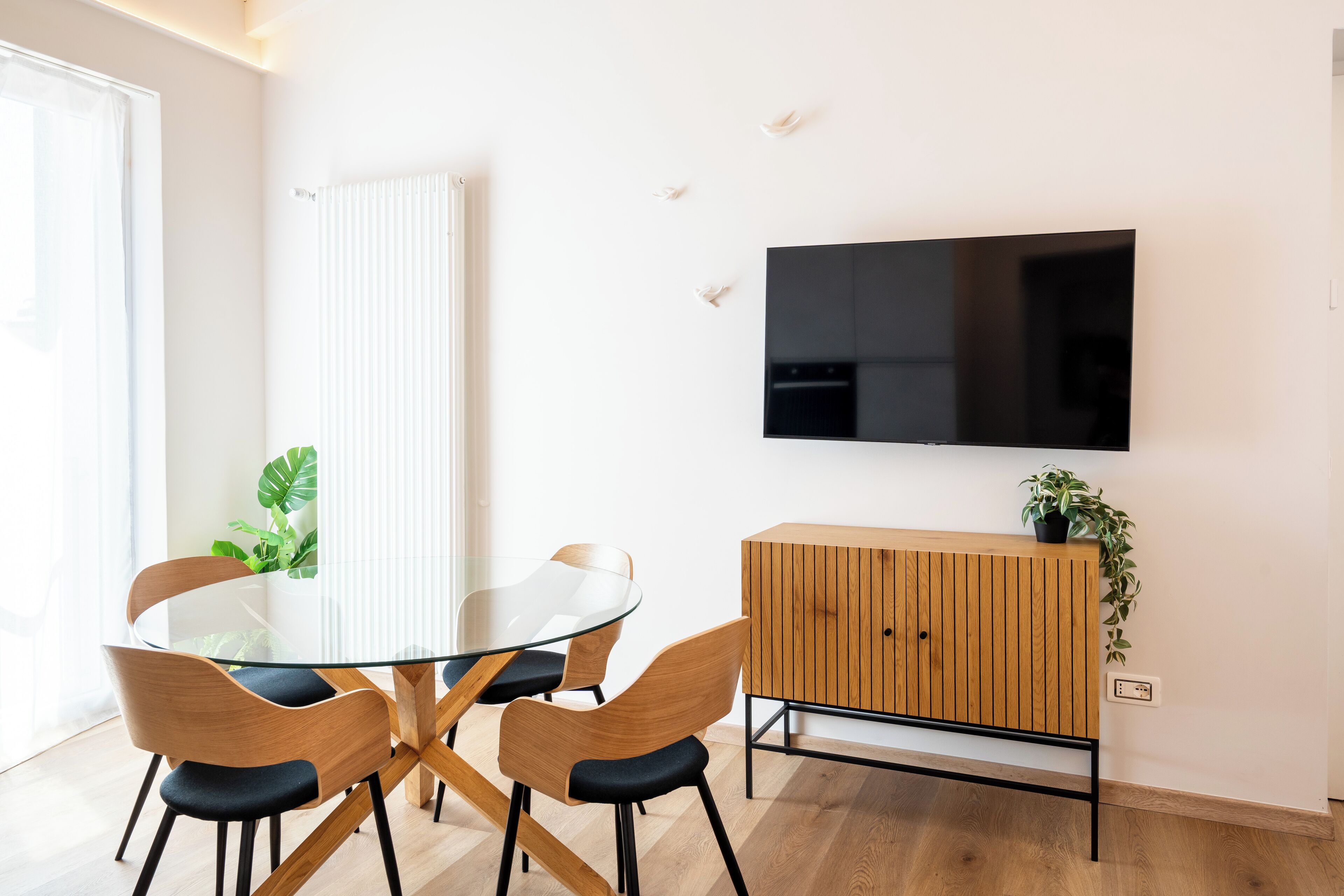 Ein moderner Essbereich mit einem Glastisch, Holzstühlen, einem Holzschrank und einem an der Wand befestigten Fernseher.