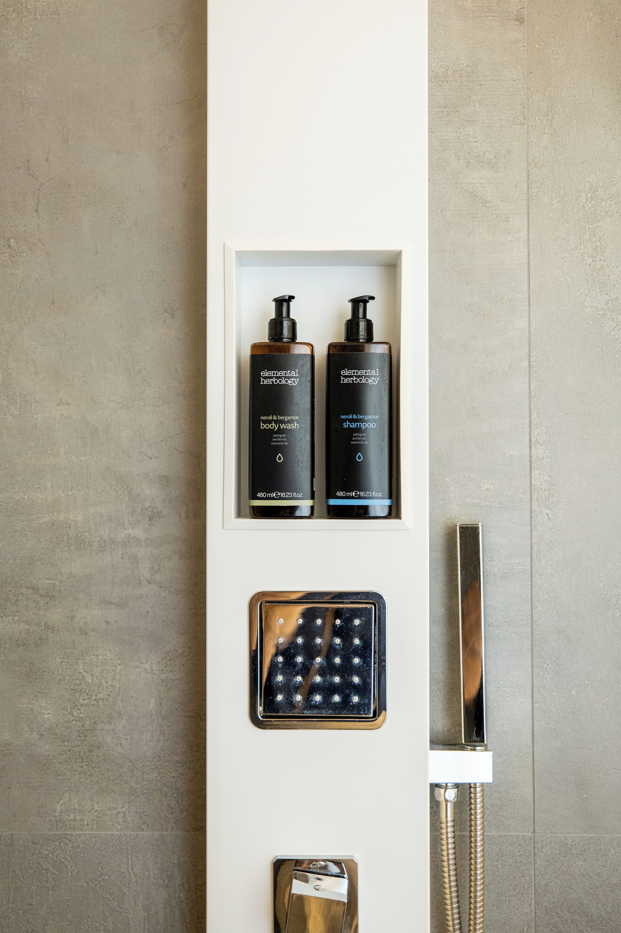 A bathroom with a shower, a framed display of two Elemental Herbology body wash bottles, and a handheld showerhead.