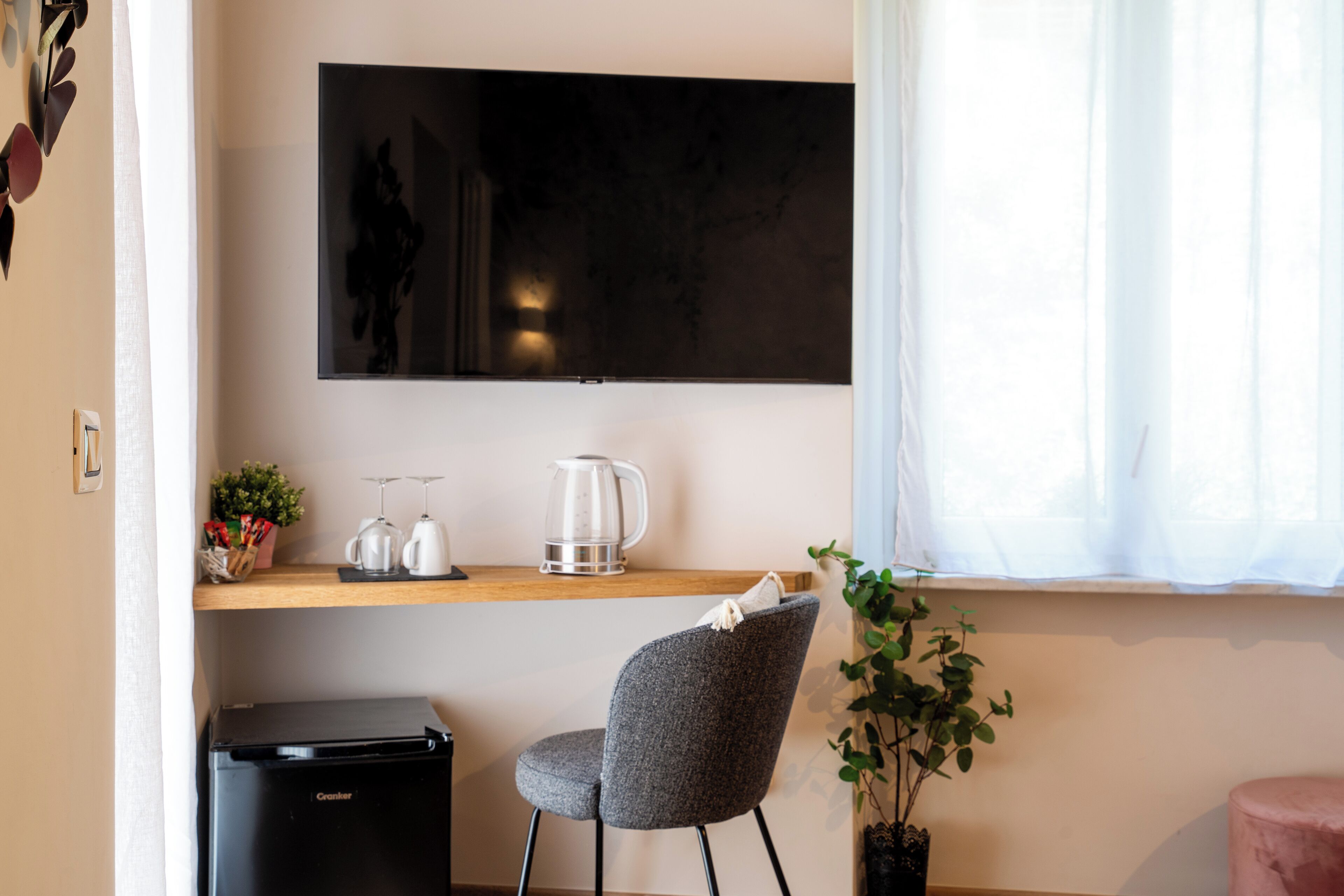 A modern room with a flat-screen TV mounted on the wall, a small kitchenette with a coffee maker, a grey chair, and a potted plant.