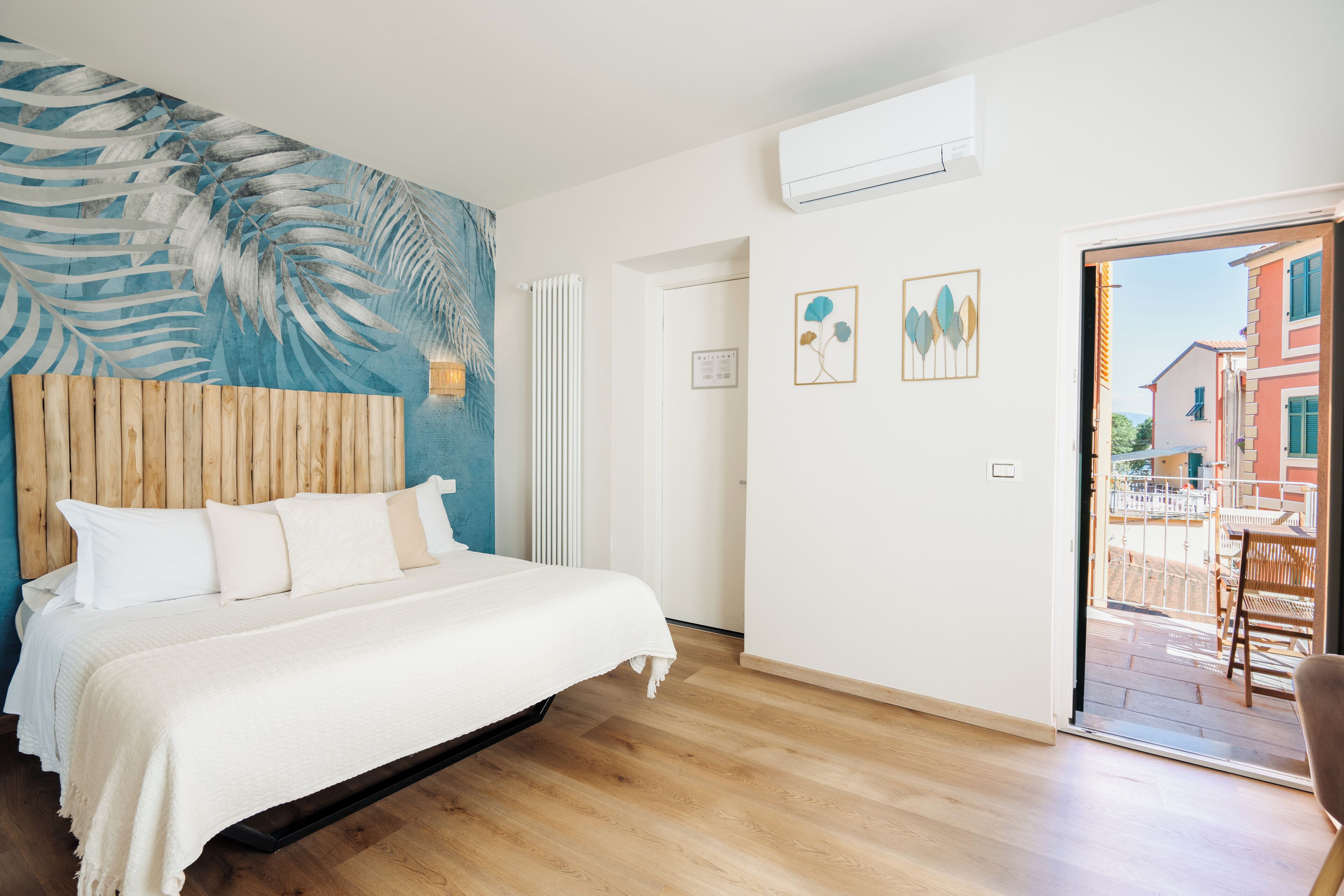 Ein Schlafzimmer mit einem Bett, einem Holzkopfbrett, einer blauen Wandmalerei, einem weißen Heizkörper und einer Tür, die zu einem Außenbereich mit Tisch und Stühlen führt.