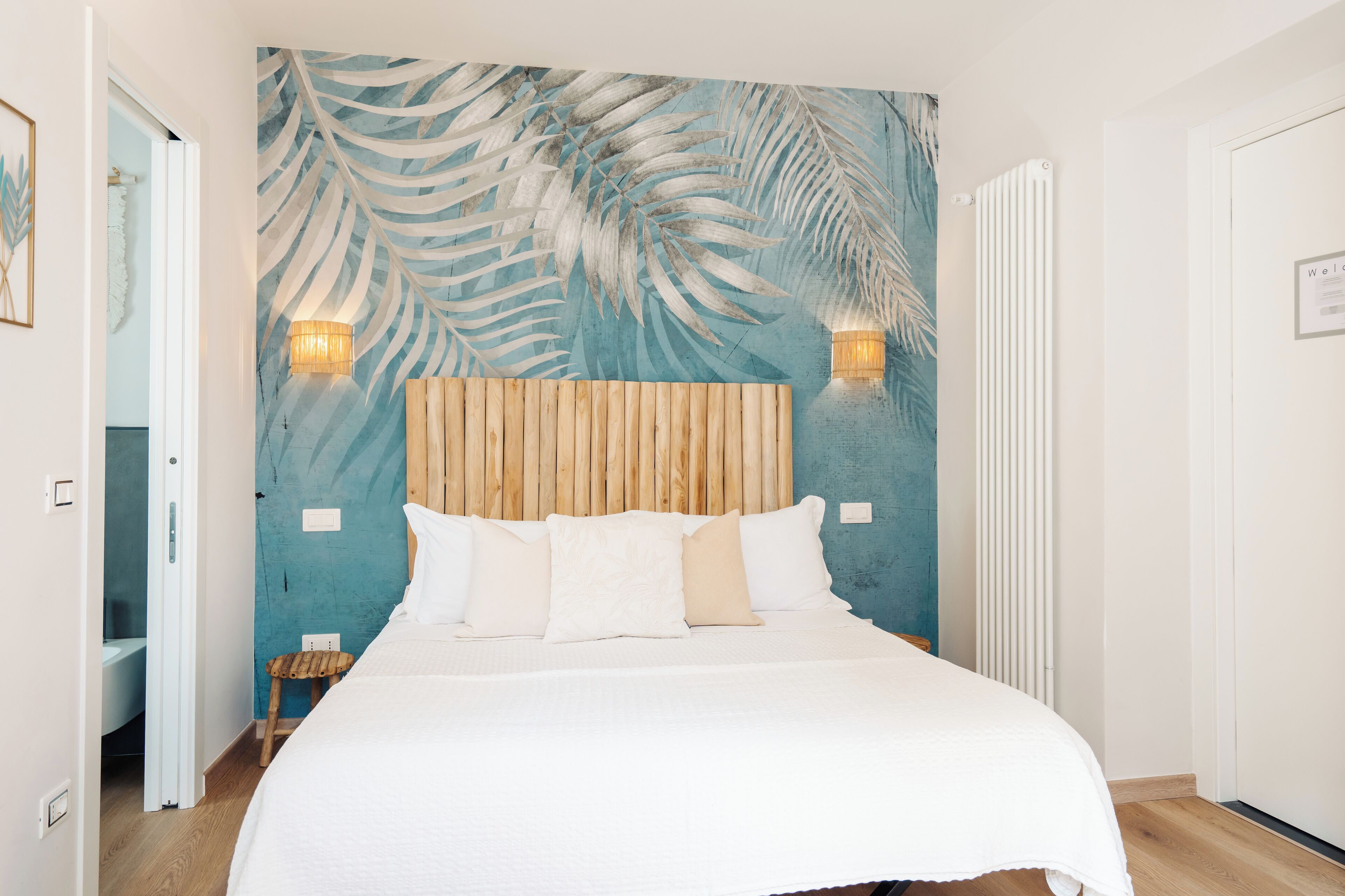 Un dormitorio con cama, cabecera de madera, dos luces fijadas a la pared y un mural con motivos de hojas tropicales.
