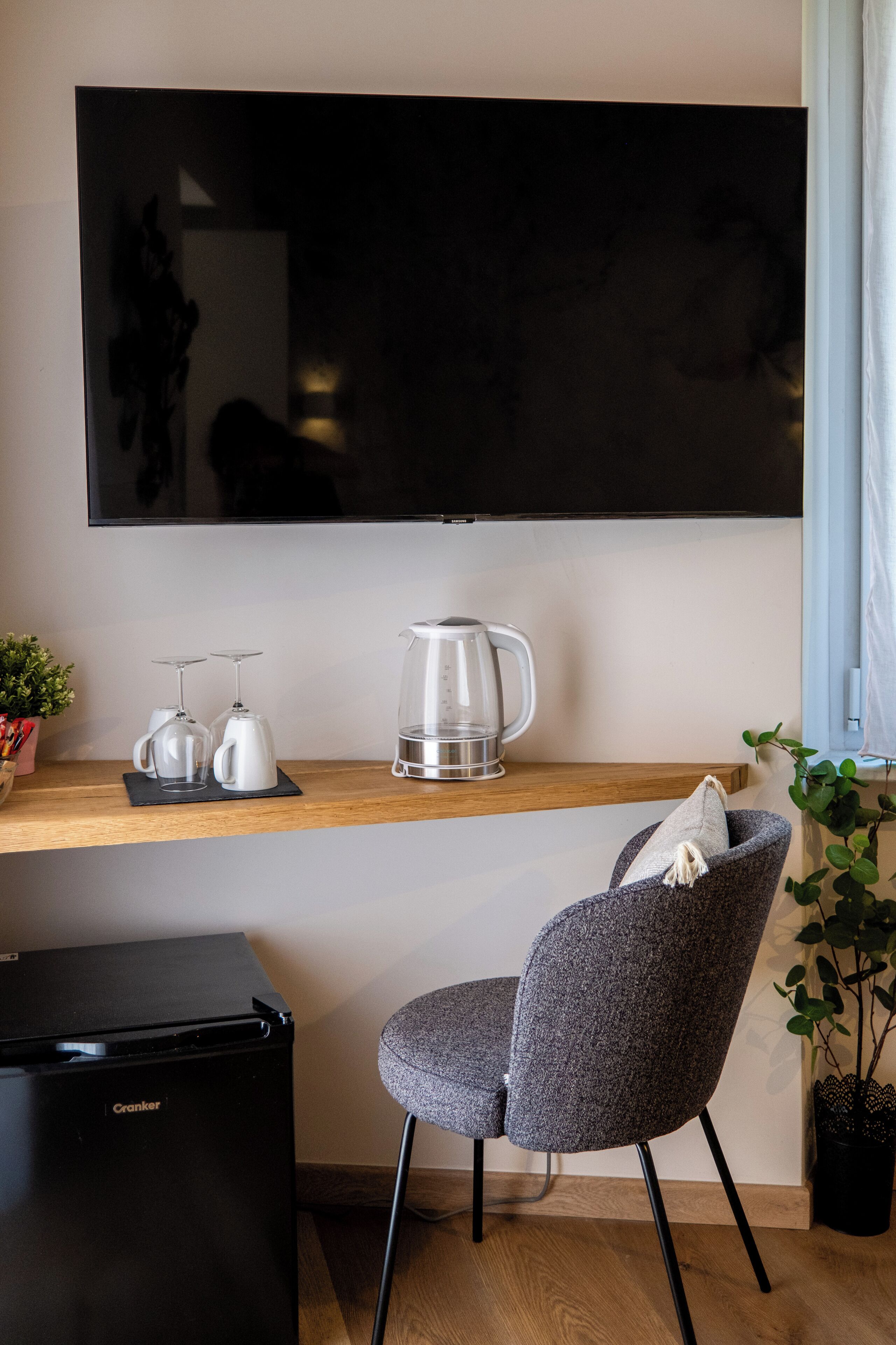 A modern living space with a flat-screen TV mounted on the wall, a coffee maker, two wine glasses, a small refrigerator, and a grey upholstered chair.