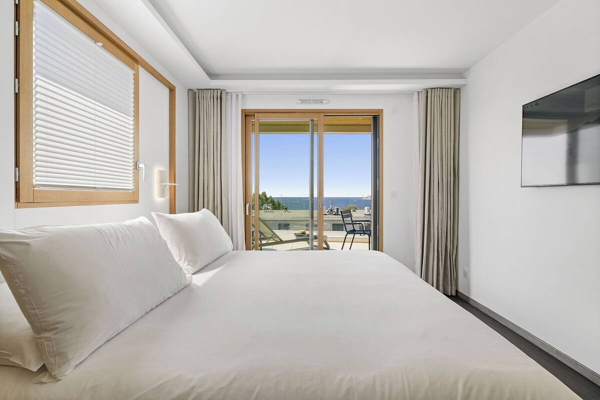 A hotel room with a large bed, a TV mounted on the wall, and a balcony with a view of the sea.