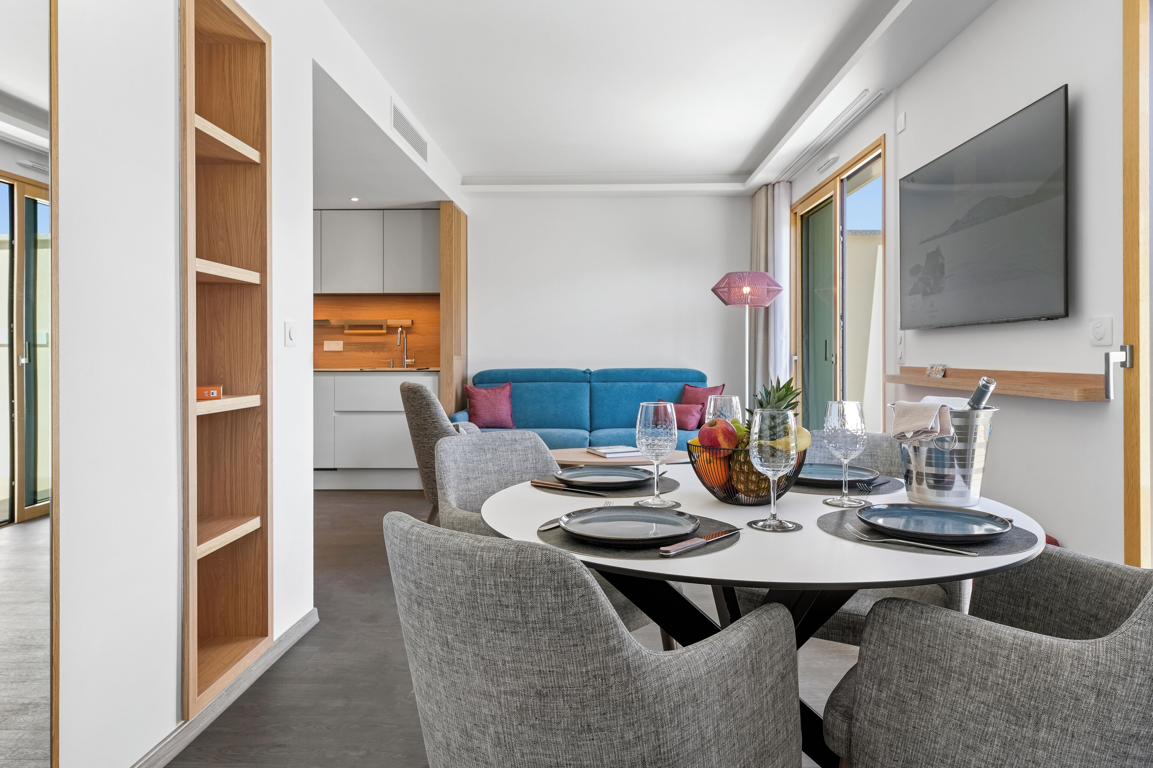 Un comedor moderno con una mesa redonda dispuesta para cuatro comensales, sillas grises tapizadas, un sofá azul al fondo y un televisor de pantalla plana grande montado en la pared.