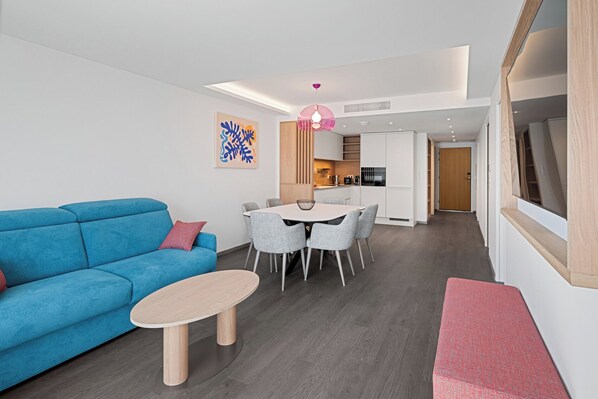A modern living room with a blue sofa, a round coffee table, and a dining area with grey chairs. - Novaya Suites (Ajaccio)