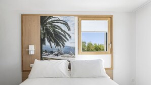 A bedroom with a bed, a window with blinds, a door with a wooden frame, and a wall with a large tropical mural.