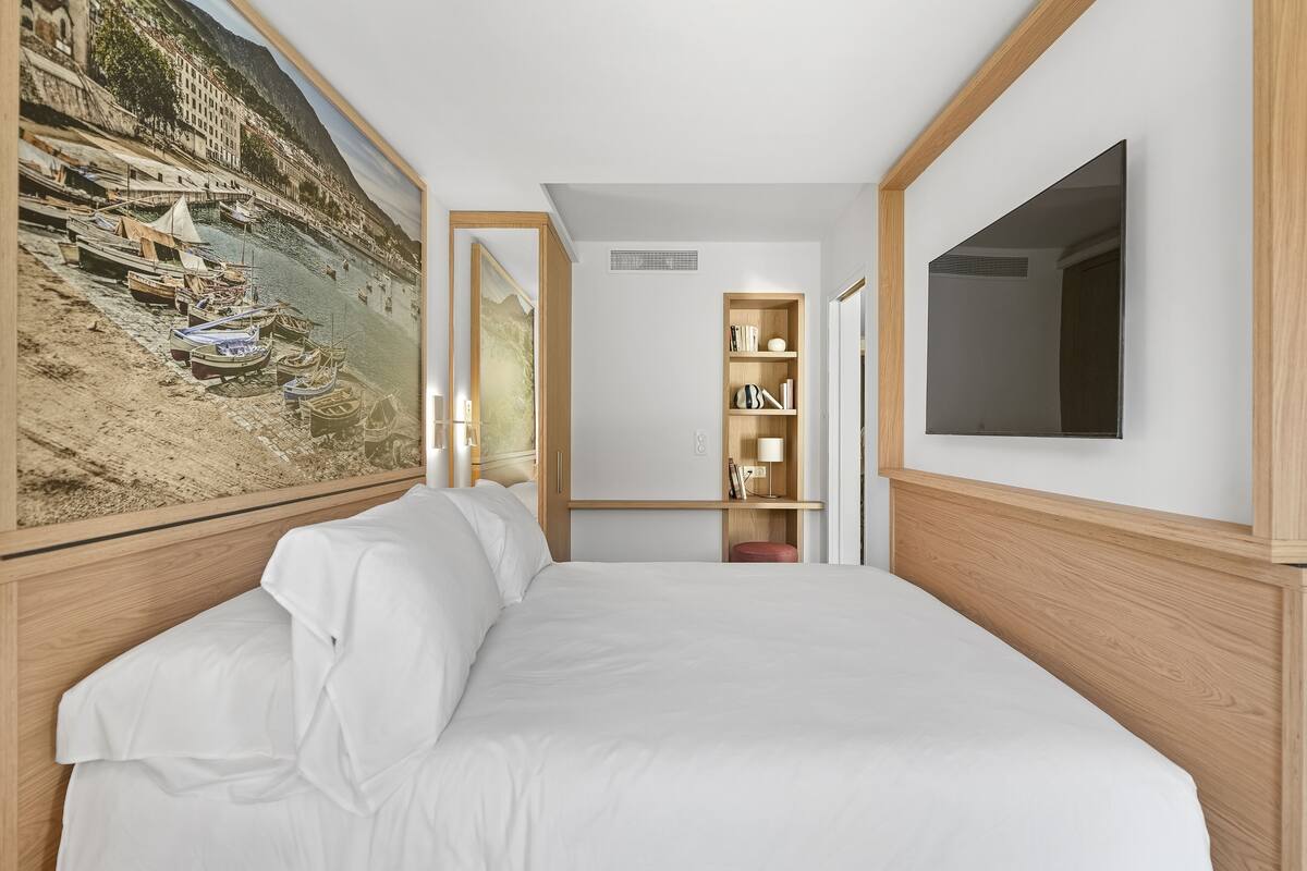 A hotel room with a large bed, a wooden headboard, a bookshelf, and a flat-screen TV mounted on the wall.
