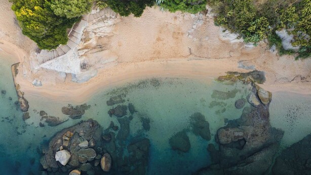 An aerial view of a beach with clear waters, rocky formations, and a staircase leading down to the shore.